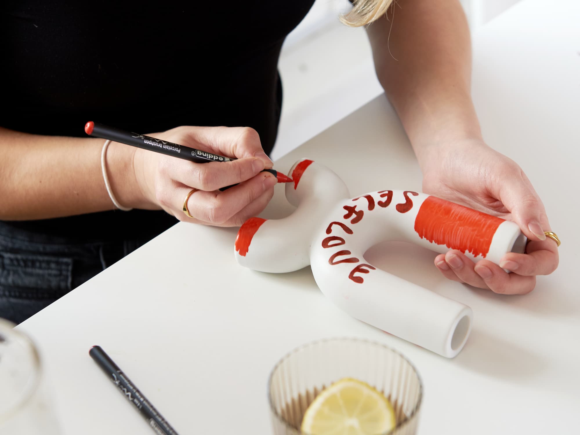 Jemand bemalt eine weiße Keramikvase mit roter Farbe und dem edding 4200 Porzellanpinselstift von edding, schreibt „SELF LOVE“ darauf und fügt farbige Details hinzu. Ein Zitronenwasserglas und ein schwarzer Marker stehen auf dem weißen Tisch – ideal zum Keramikdekorieren.