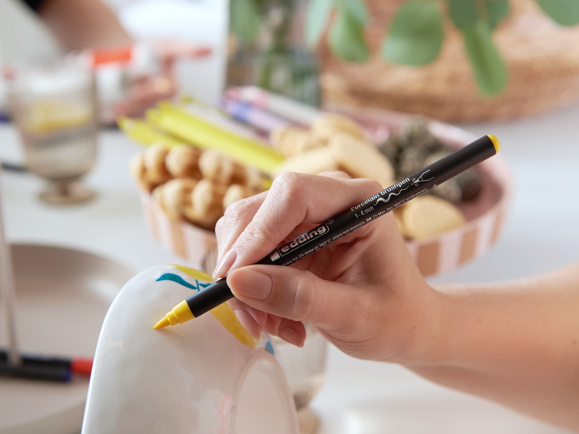 Eine Hand verwendet den edding 4200 Porzellanpinselstift von edding mit gelber Spitze, um einen weißen Keramikgegenstand zu dekorieren. Keramik dekorieren ist dadurch einfach und spülmaschinenfest. Eine Snackschale und Grünpflanzen sind im Hintergrund verschwommen.