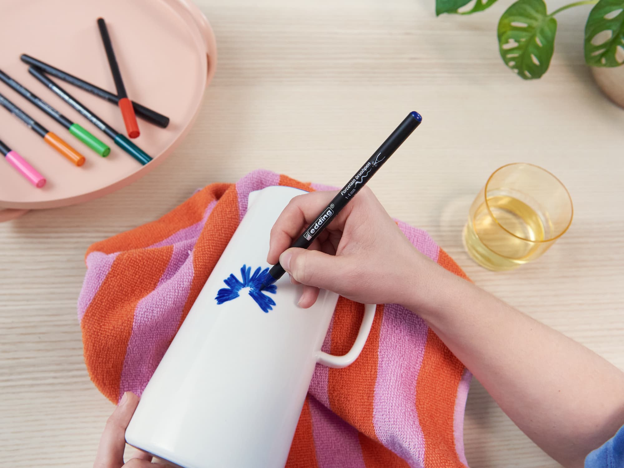 Eine Person verziert eine weiße Tasse mit einer blauen Blume. Dazu verwendet sie den edding 4200 Porzellanpinselstift von edding und hält sie über ein orange-rosa gestreiftes Handtuch. In der Nähe stehen auf dem hellen Holztisch Keramik-Dekorationsutensilien, ein Getränk und eine Pflanze.