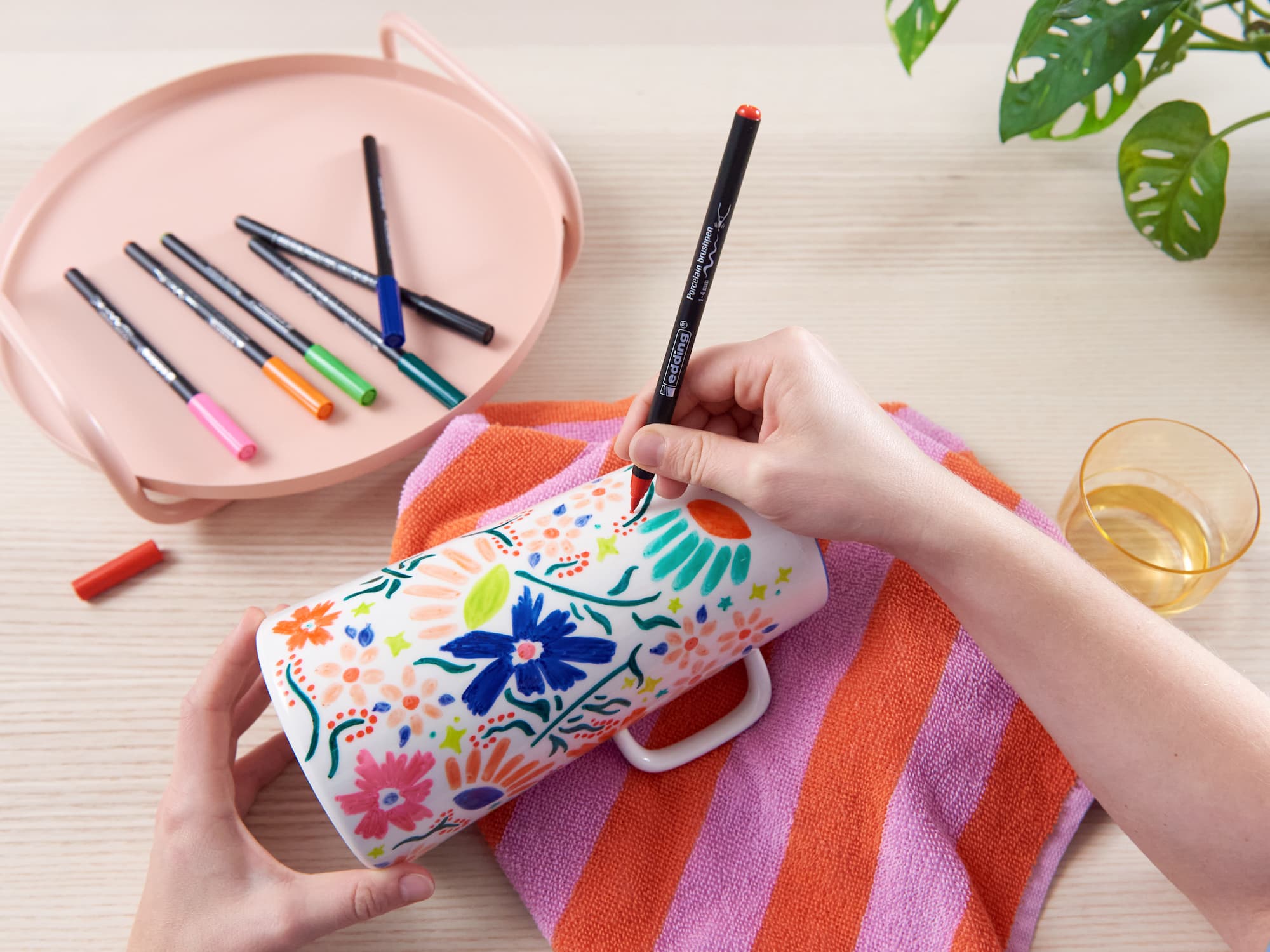 Eine Person verziert eine weiße Tasse mit bunten Blumenmustern und verwendet dazu edding 4200 Porzellanpinselstifte von edding. Die Tasse steht auf einem rosa-orange gestreiften Handtuch. Auf dem hellen Holztisch stehen weitere Stifte, ein Tablett, ein Glas und eine grüne Pflanze.