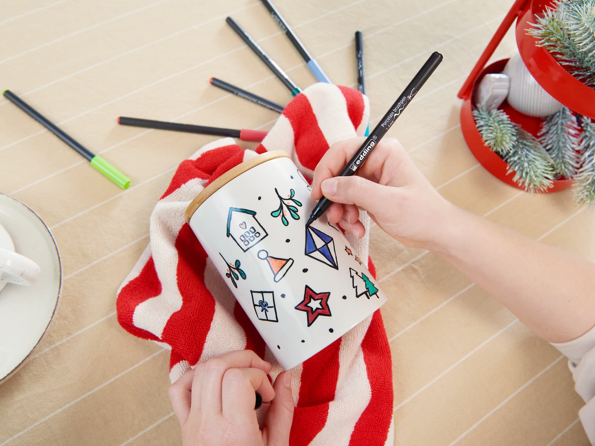 Mit einem edding 4200 Porzellanpinselstift von edding verziert jemand ein weißes Keramikgefäß mit weihnachtlichen Motiven auf einem rot-weiß gestreiften Tuch, umgeben von weiteren Markern und festlichem Dekor auf der beige gestreiften Oberfläche.