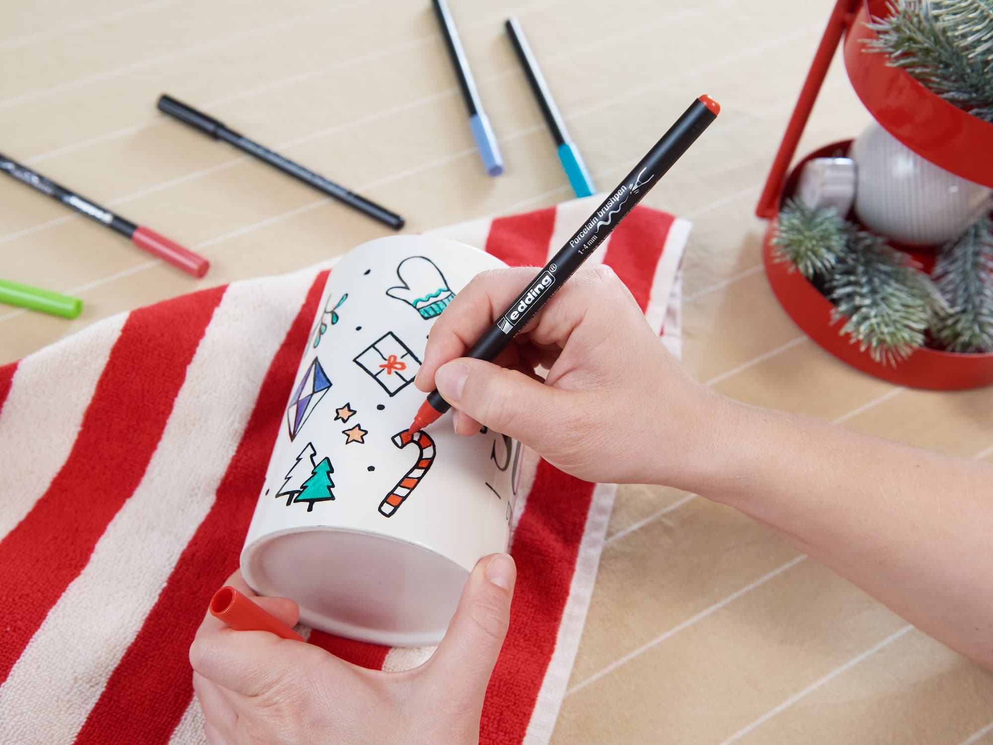 Jemand verwendet den edding 4200 Porzellanpinselstift von edding, um weihnachtliche Motive wie eine Zuckerstange und einen Weihnachtsbaum auf eine weiße Tasse zu malen. Farbige Marker, ein rotes Handtuch und festliche Dekoration stehen für „Keramik dekorieren“ auf dem Tisch.