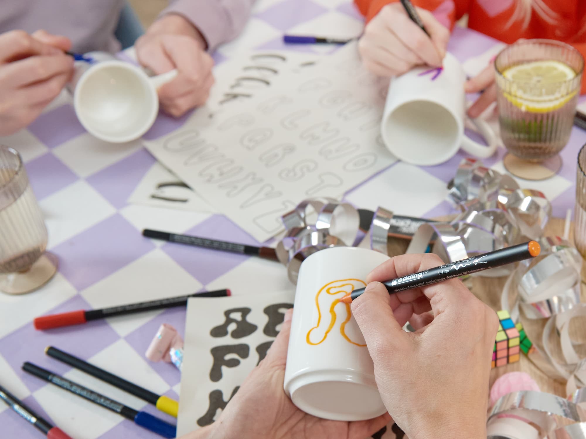 Drei Personen verwenden den edding 4200 Porzellanpinselstift von edding zum Dekorieren von Keramik. Sie zeichnen einen gelben Kringel auf eine Tasse, während andere an einem Tisch mit Schablonen, Künstlerbedarf und Ausstechformen Buchstaben nachzeichnen.