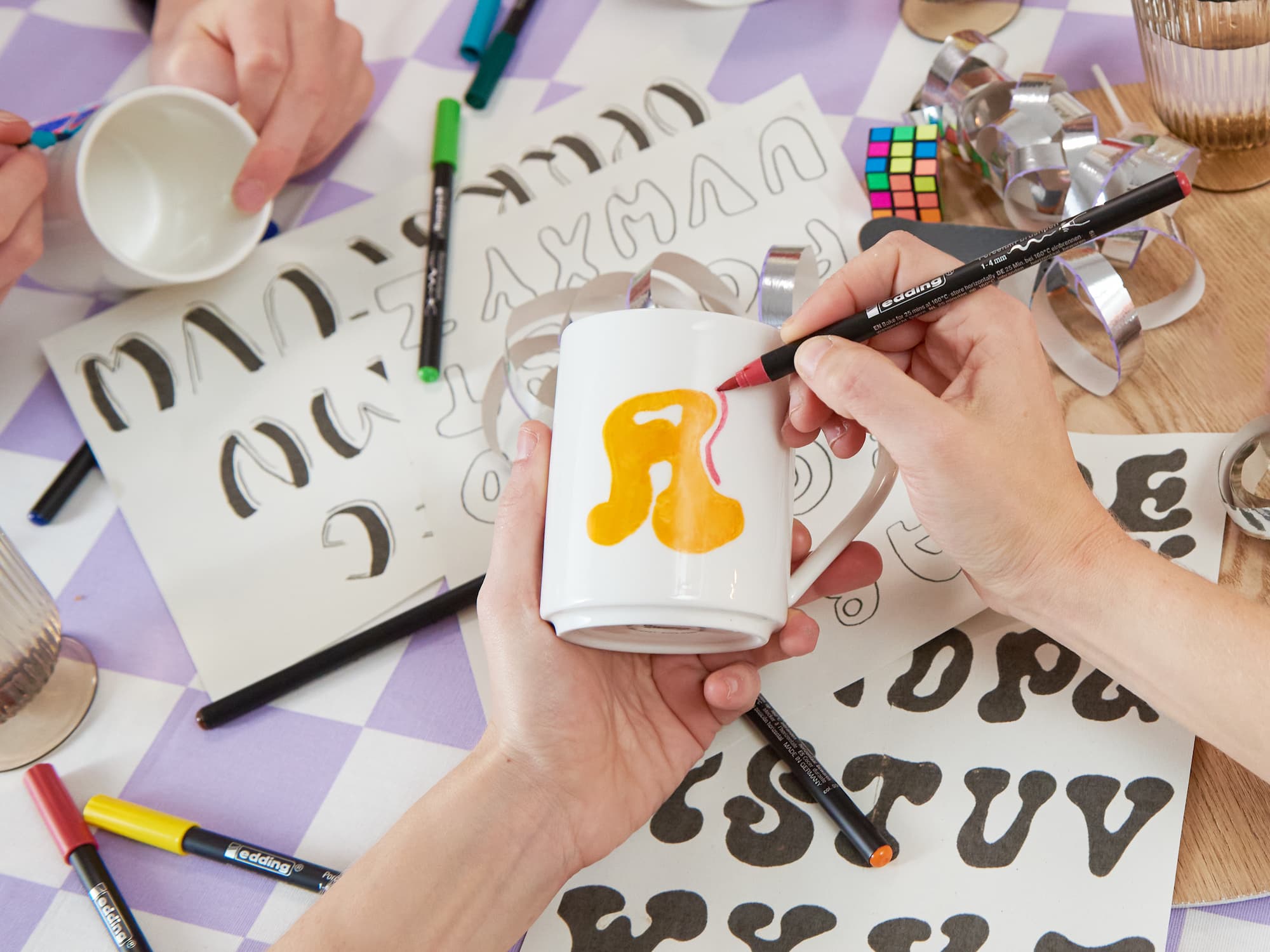 Hände verzieren eine weiße Tasse mit einem gelb-rosa „R“ mit dem edding 4200 Porzellanpinselstift von edding. Daneben liegen Marker, Tassen, fettgedruckte Alphabetblätter, ein Zauberwürfel und ein Metallband – ideal zum Dekorieren von Keramik.