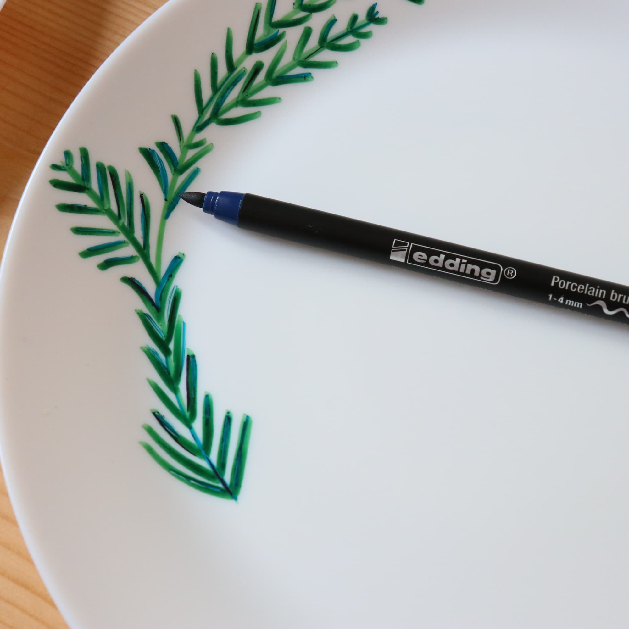 Der edding 4200 Porzellanpinselstift von edding zeichnet grüne, blattähnliche Muster auf weißem Teller – spülmaschinenfest und ideal, um Keramik stilvoll zu dekorieren.