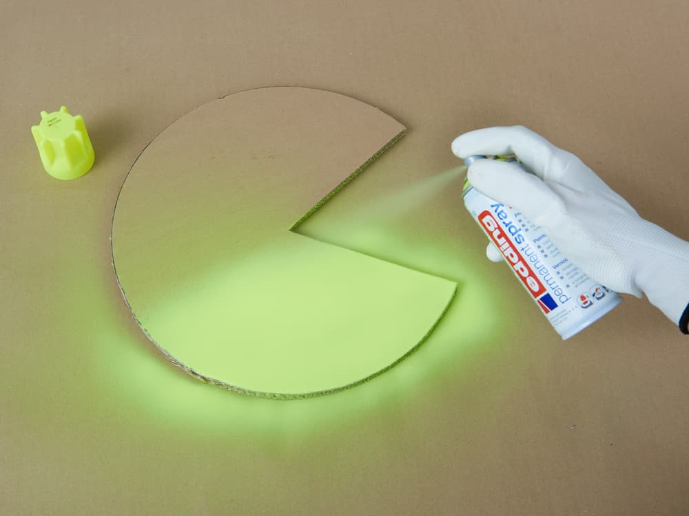 Eine Hand in einem weißen Handschuh trägt Edding 5200 Permanentspray Acryllack Neon & kräftige Farben aus einer Tube auf ein rundes Stück grünes Material auf, aus dem ein kuchenförmiger Abschnitt entfernt wurde, auf einer braunen Oberfläche, daneben liegt ein neonfarbener Textmarker.