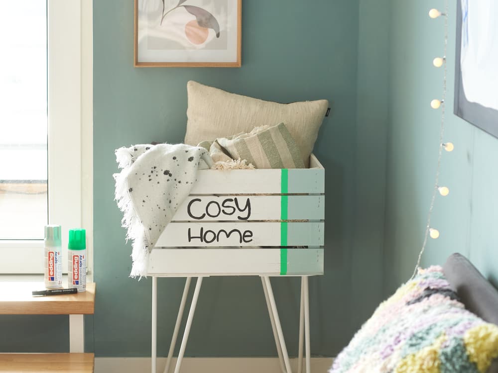 Eine gemütliche Ecke mit einer weißen Kiste mit der Aufschrift „Cosy Home“, geschmückt mit weichen Kissen und einer gepunkteten Decke, steht neben einer Reihe warmer Lichterketten und schafft eine beruhigende Atmosphäre mit edding 5200 Permanentspray Acryllack Neon & kräftige Farben.