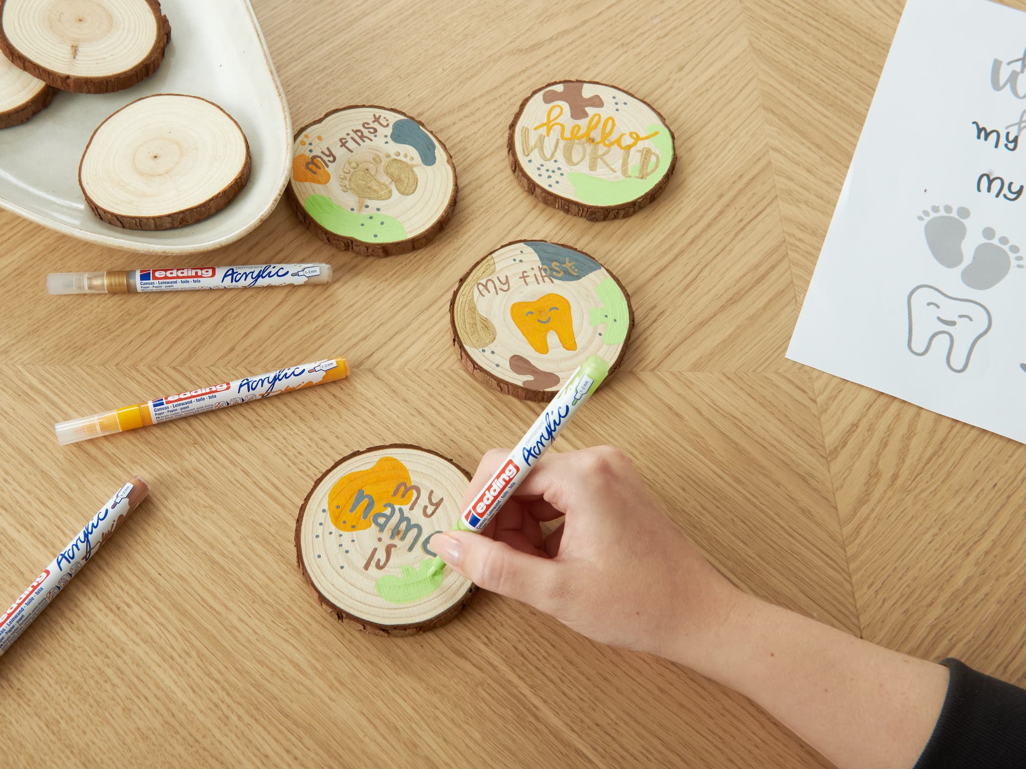 Die Hand einer Person hält einen edding Acrylmarker und dekoriert Holzuntersetzer mit bunten Zeichnungen und handgeschriebenen Sätzen wie „mein erstes Lächeln“.