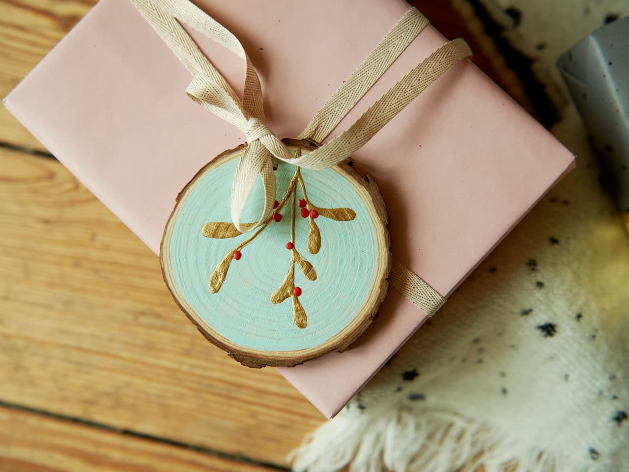 Ein hübsch verpacktes rosa Geschenk mit einem rustikalen Holzornament, verziert mit einer bemalten Mistel mit edding 5000 Acrylmarker breit 5er-Set, zusammengebunden mit einem cremefarbenen Band, ruht auf einer Holzoberfläche neben einem gemütlichen