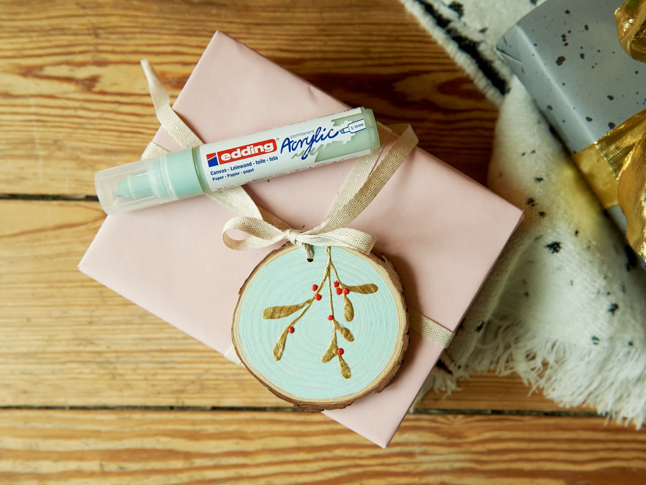Ein wunderschön verpacktes Geschenk, verziert mit einem handbemalten Holzornament und einem darauf platzierten edding 5000 Acrylmarker breit 5er-Set, das einem festlichen Geschenk eine persönliche Note verleiht.