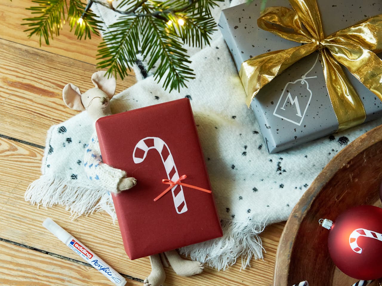 Festliche Weihnachtsgeschenke verpackt in elegantem Papier mit Schleifen, ein kuscheliger Schal, saisonale Dekorationen, hervorgehoben durch edding 5100 Acrylmarker-Medium-Designs, und ein Hauch von Grün auf einem Holztisch, der die Wärme einfängt.