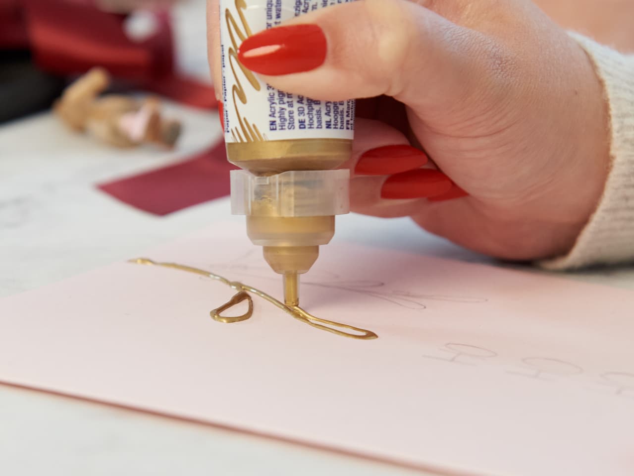 Eine Person, die mit Präzision bastelt und eine dünne Linie goldenen Glitzerklebers aus einem edding 5400 Acryl 3D Double Liner auf rosa Papier drückt.