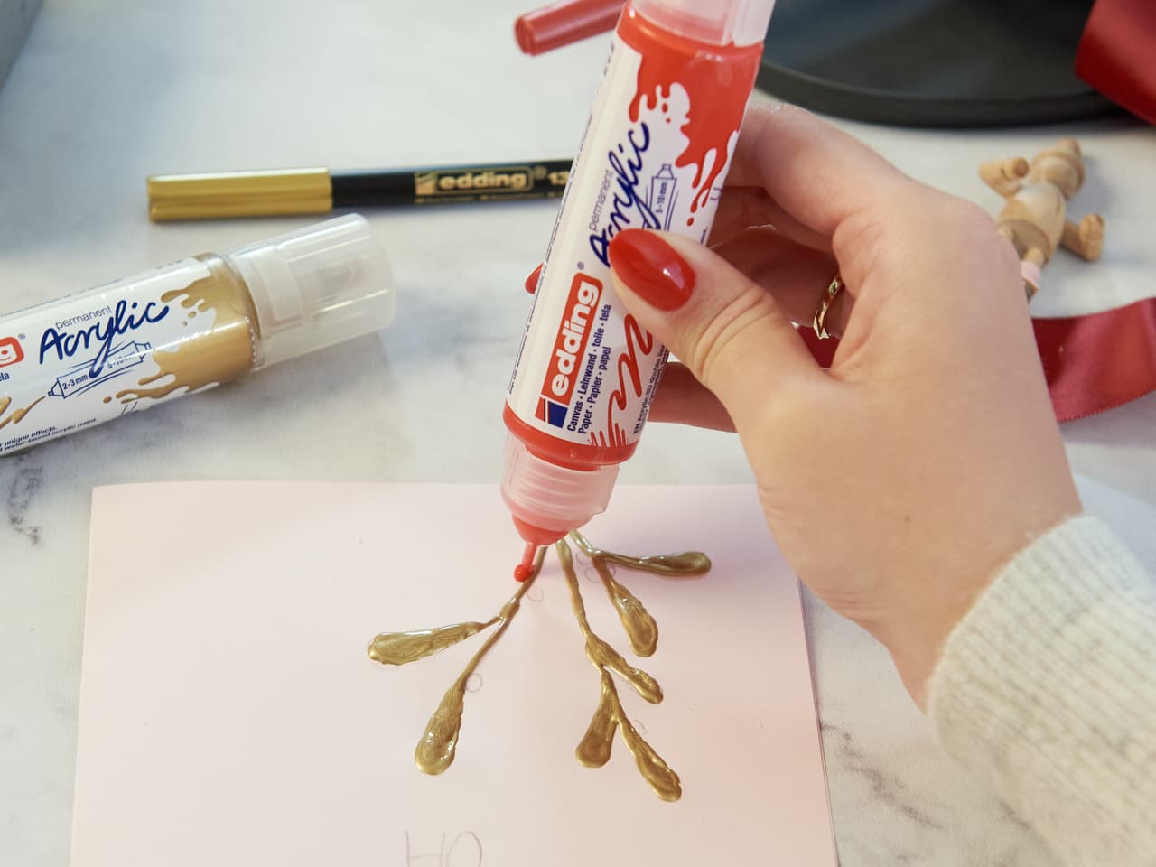 Eine Hand drückt eine Tube roten edding 5400 Acryl 3D Double Liner auf ein Blatt Papier mit goldenem Farbdesign, in der Nähe befinden sich andere Künstlerutensilien.