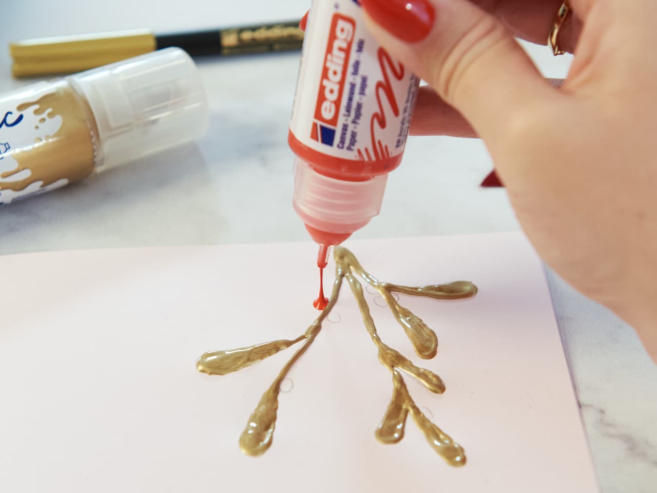 Eine Person, die Kunst mit einem edding 5400 Acryl 3D Double Liner 5er-Set mit roter Spitze kreiert und eine goldene, hochwertige edding Acrylfarbe in einem verzweigten Muster auf Papier aufträgt