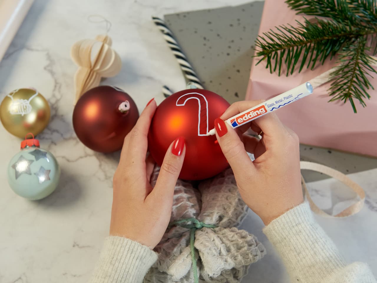 Hände mit rotem Nagellack personalisieren einen Weihnachtsschmuck mit einem edding 751 Glanzlackmarker, umgeben von festlichen Dekorationen.