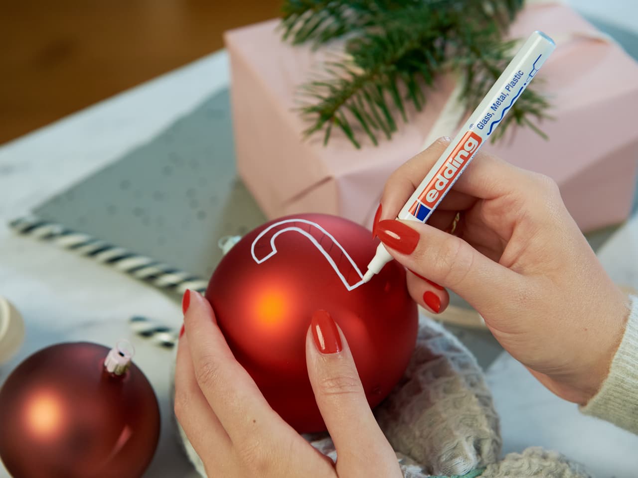Weihnachtsstimmung basteln: Eine Person verziert einen roten Weihnachtsschmuck mit einem Glanzlackmarker edding 751 und verleiht so festlicher Dekoration eine persönliche Note.