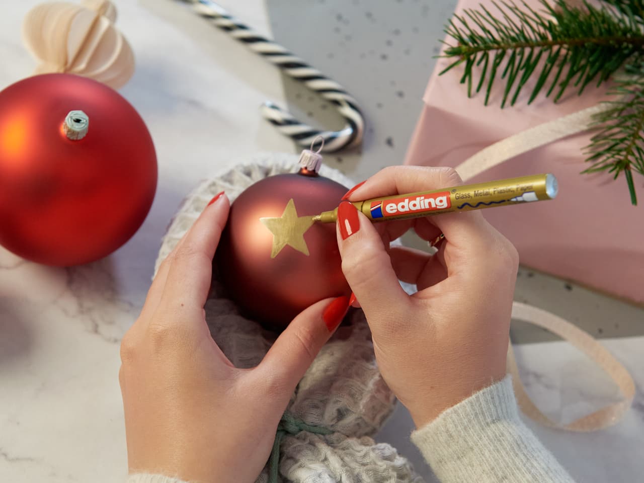 Eine Person verziert mit einem edding 751 Glanzlackmarker einen roten Weihnachtsschmuck mit einem goldenen Stern, im Hintergrund Weihnachtsdekoration und ein Geschenk.