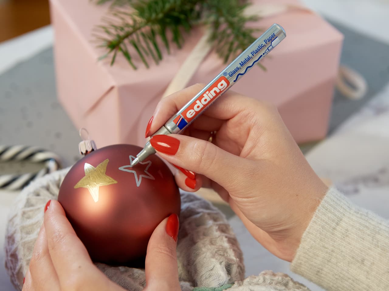 Hände dekorieren mit einem Glanz-Lackmarker edding 751 zart eine rote Weihnachtskugel mit einem silbernen Stern, im Hintergrund Weihnachtsgeschenke und Grünpflanzen.