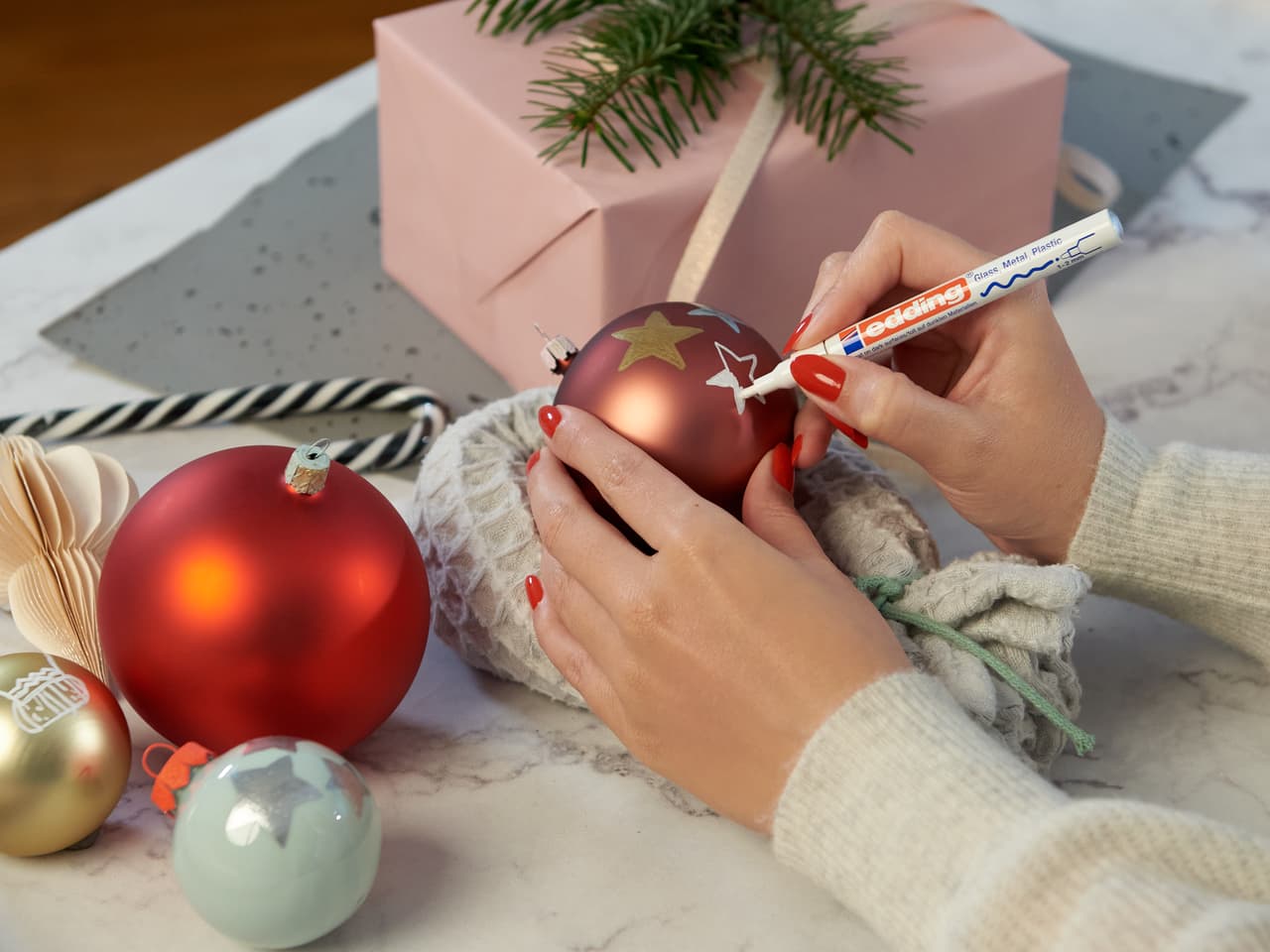 Personalisieren Sie einen Weihnachtsschmuck: Hände dekorieren sorgfältig eine festliche rote Kugel mit einem edding 751 Glanzlackmarker 3er-Set auf Glasoberflächen, mit zusätzlicher Weihnachtsdekoration und einer Geschenkbox in der Nähe.