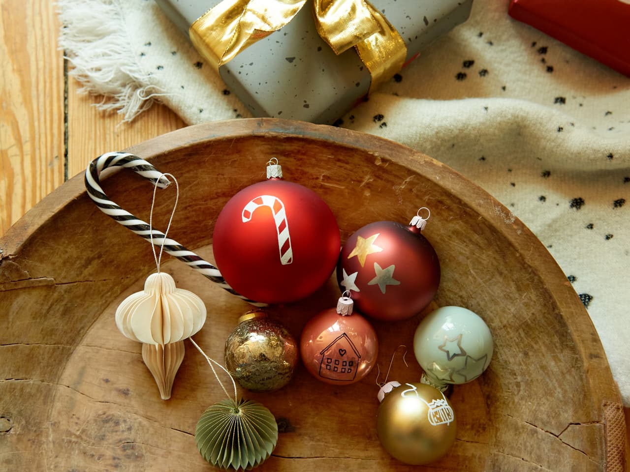 Eine festliche Reihe von Weihnachtsornamenten und einer goldenen Geschenkbox auf einer rustikalen Holzoberfläche, die zum Verschenken und Dekorieren an Feiertagen gedacht ist, mit Beschriftungen, die mit dem edding 751 Glanzlackmarker 3er-Set für Glasoberfl erstellt wurden.