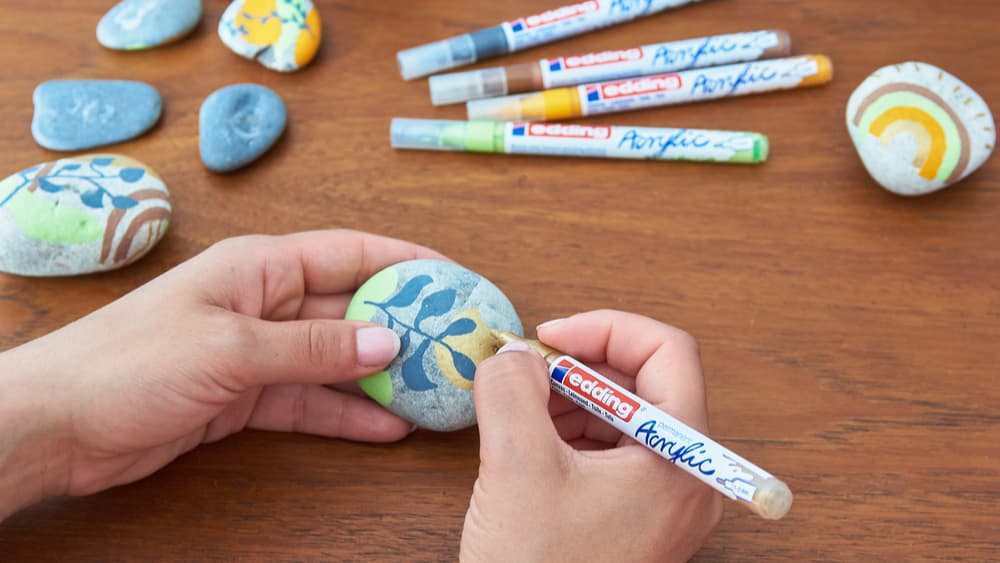 Eine Person zeichnet mit einem Acrylfarbenmarker Blätter auf einen glatten Stein. Bemalte Steine und Marker in verschiedenen Farben liegen verstreut auf einem Holztisch.