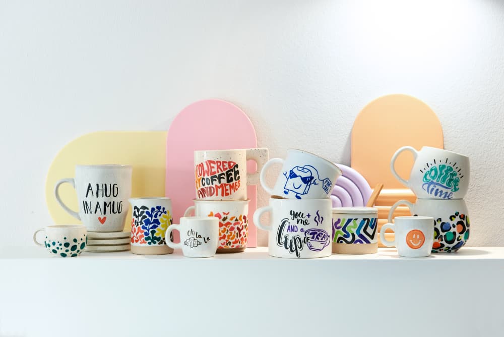 The edding 4200 Porzellanpinselstift 6er-Set offers cheerful patterns and playful text for those who enjoy Keramik bemalen. These mugs are displayed on a white shelf with a vibrant, abstract background in pink, purple, and orange.