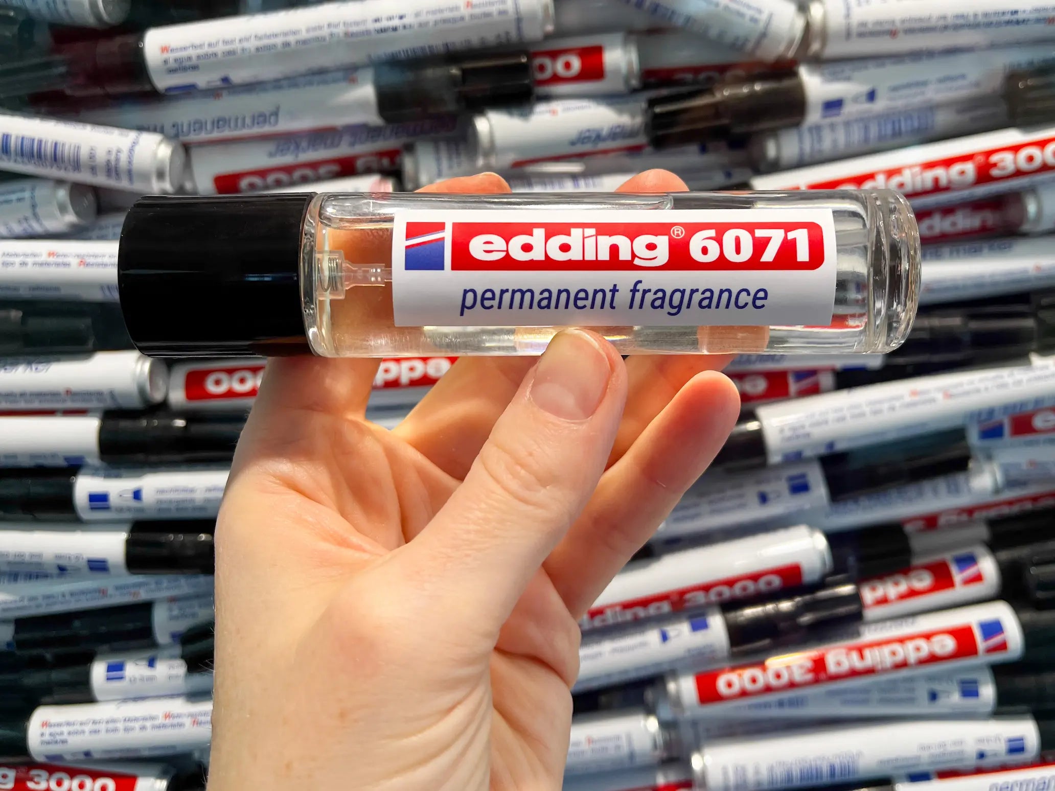 Eine Hand hält eine klare Flasche mit der Aufschrift "edding 6071 permanent fragrance" von edding, im Hintergrund sind edding Marker zu sehen.