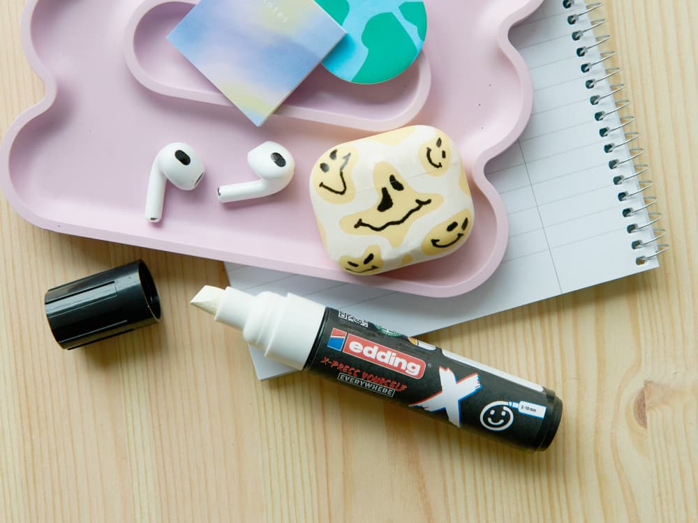 Eine Tischplatte mit einem rosa Tablett, auf dem ein Paar AirPods, ein kleiner Untersetzer mit Smiley-Motiv und bunte Zettel liegen. Unter dem Tablett liegt ein abgenommener edding 6900 X Multimarker aus dem Black & White Color 2er-Set von edding neben einem aufgeschlagenen linierten Notizbuch.