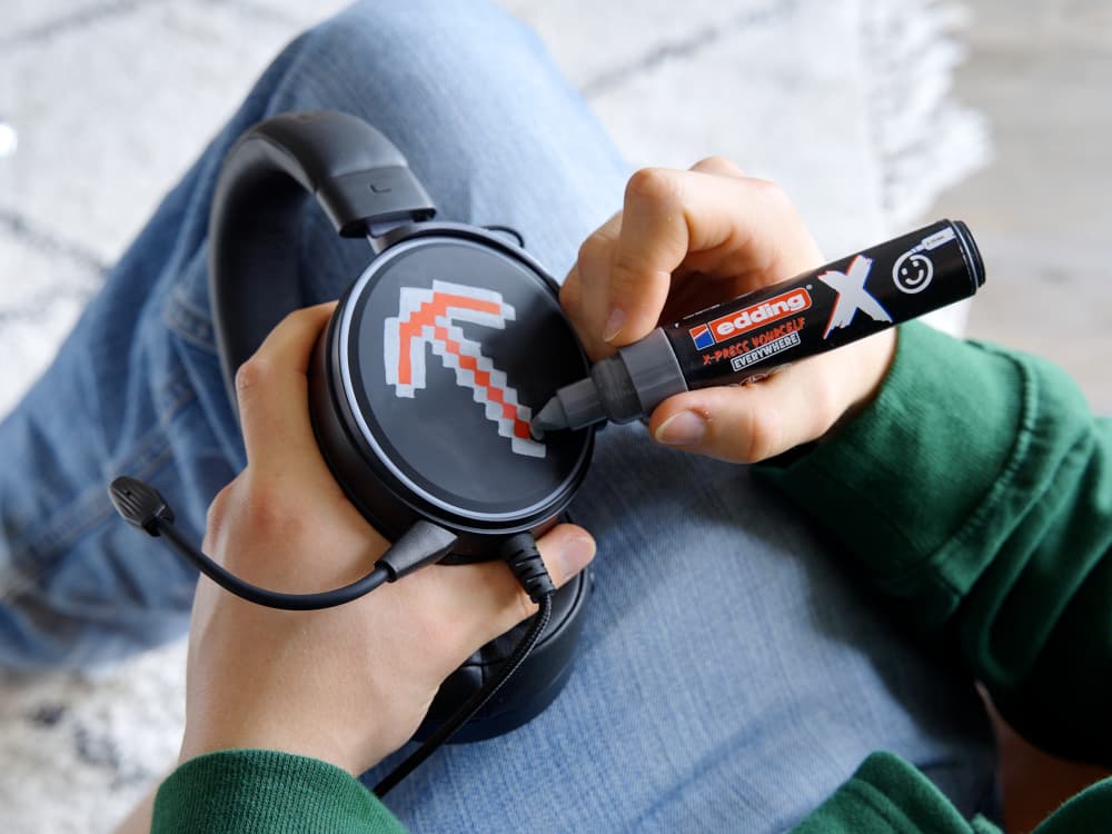 Eine Person im grünen Hemd zeichnet mit einem edding 6900 X Multimarker Yay Grey Color aus dem 2er-Set eine rot-weiße, pixelige Spitzhacke auf die Seite eines schwarzen Over-Ear-Headsets mit Mikrofon. Mit einer Hand hält die Person das Headset fest, während sie mit der anderen gekonnt die Striche des vielseitigen Markers aufträgt.