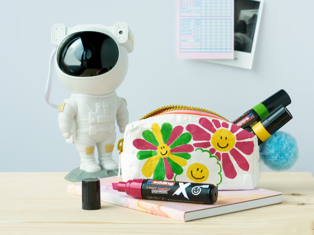 Auf einem Schreibtisch steht ein buntes Federmäppchen mit lächelnden Blumenmotiven neben Markern, darunter ein edding 6900 X Multimarker Candy Color 3er-Set mit langlebiger Tinte. Dahinter steht eine kleine Astronautenfigur. Am Federmäppchen ist ein hellblauer Pompon befestigt, im Hintergrund hängen ein Planer und ein Foto an der Wand.