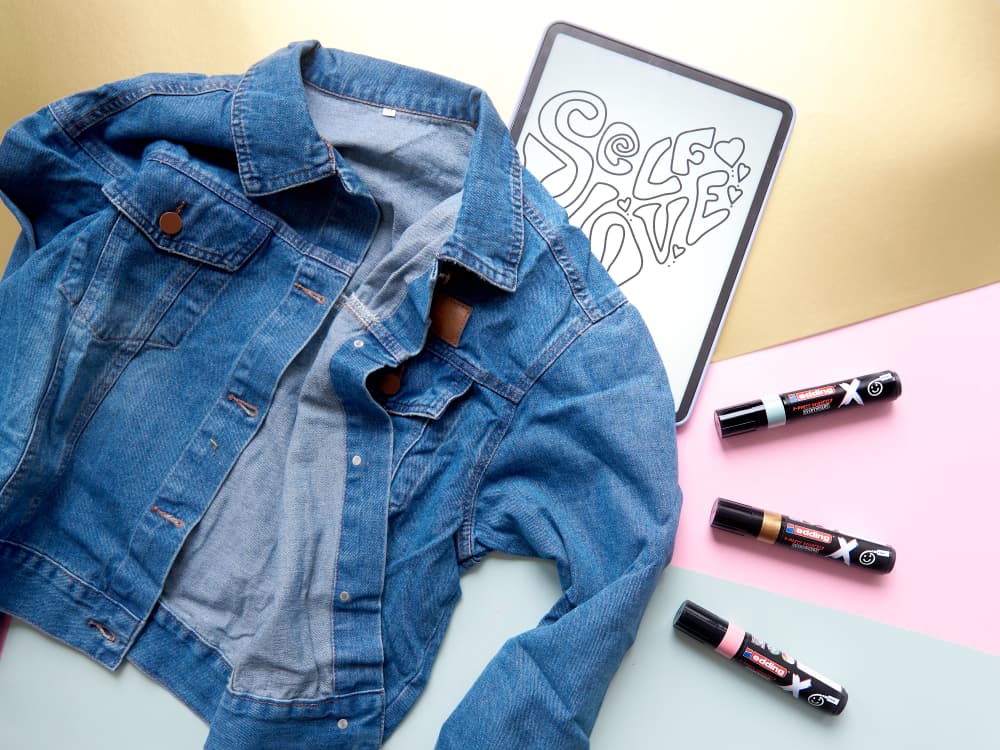 Eine Jeansjacke liegt offen auf einem pastellfarbenen Untergrund neben einem aufgeschlagenen Skizzenbuch, in das mit dem edding 6900 X Multimarker Pastel Color 3er-Set in kräftigen, dekorativen Buchstaben das Wort „SELFIE“ geschrieben ist. Neben dem Skizzenbuch liegen drei Lippenstifthülsen mit schwarz-weißer Verpackung.