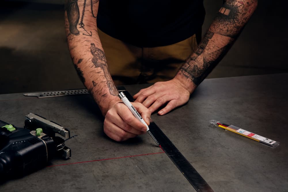 Eine Person mit tätowierten Armen zeichnet mit Lineal und Stift eine rote Linie auf eine Metalloberfläche, um präzise Markierungen zu setzen. Auf der Werkbank liegen verschiedene Werkzeuge, darunter ein Messwerkzeug und edding 8881 Baumarker Nachfüllmine farblich sortiert 6 Stück. Die Szene ist gut beleuchtet und hat einen dunklen Hintergrund.