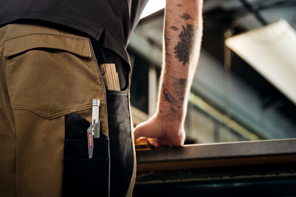 Nahaufnahme der Gesäßtasche einer Person, in der sich ein Stift und ein kleines Notizbuch befinden, darunter etwas, das wie ein edding 8880 Tiefloch Baumarker Set, inkl. Nachfüllminen Graphit aussieht. Die Person in hellbraunen und schwarzen Hosen lehnt über einer Werkbank, wobei ihr tätowierter Arm sichtbar ist. Industrielle Elemente und diffuses Licht füllen den Hintergrund.