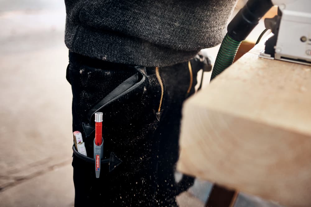 Eine Person in schwarzer Hose ist bei der Arbeit mit Holzbearbeitungsgeräten zu sehen. Werkzeuge, darunter ein roter Edding 8890 Automatischer Zimmermannsbleistift HB und ein kleiner Gegenstand, der einem Klebestift ähnelt, sind in einem an der Hose befestigten Halter verstaut. Sägespäne sind auf der Hose und in der Luft sichtbar.