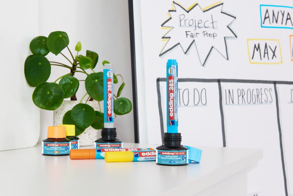 Ein edding FETK 25 Nachfülltinte Flipchart-Effektmarker und ein edding 385 stehen mit Tintennachfüllungen auf einem weißen Tisch vor einem Whiteboard mit der Aufschrift „Projekt“, „Messevorbereitung“ und Namen, ergänzt durch eine Topfpflanze für grüne Eleganz, bereit zum Hervorheben oder Nachfüllen.