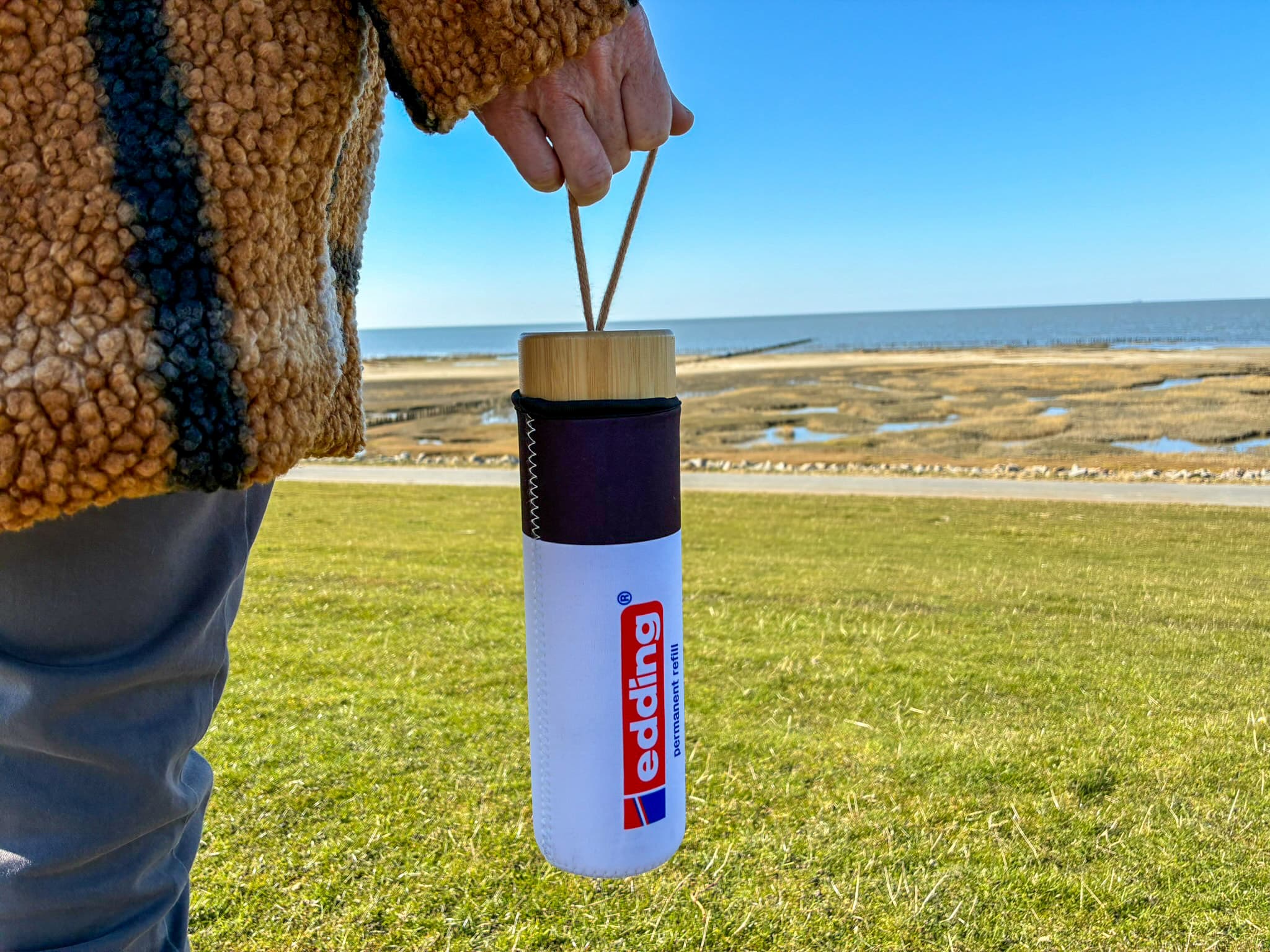 Eine Person in einem braunen Mantel hält eine edding-Glasflasche (650 ml) mit Deckel in Bambusoptik und Seilgriff und steht unter einem klaren blauen Himmel auf einer Grasfläche in Küstennähe.