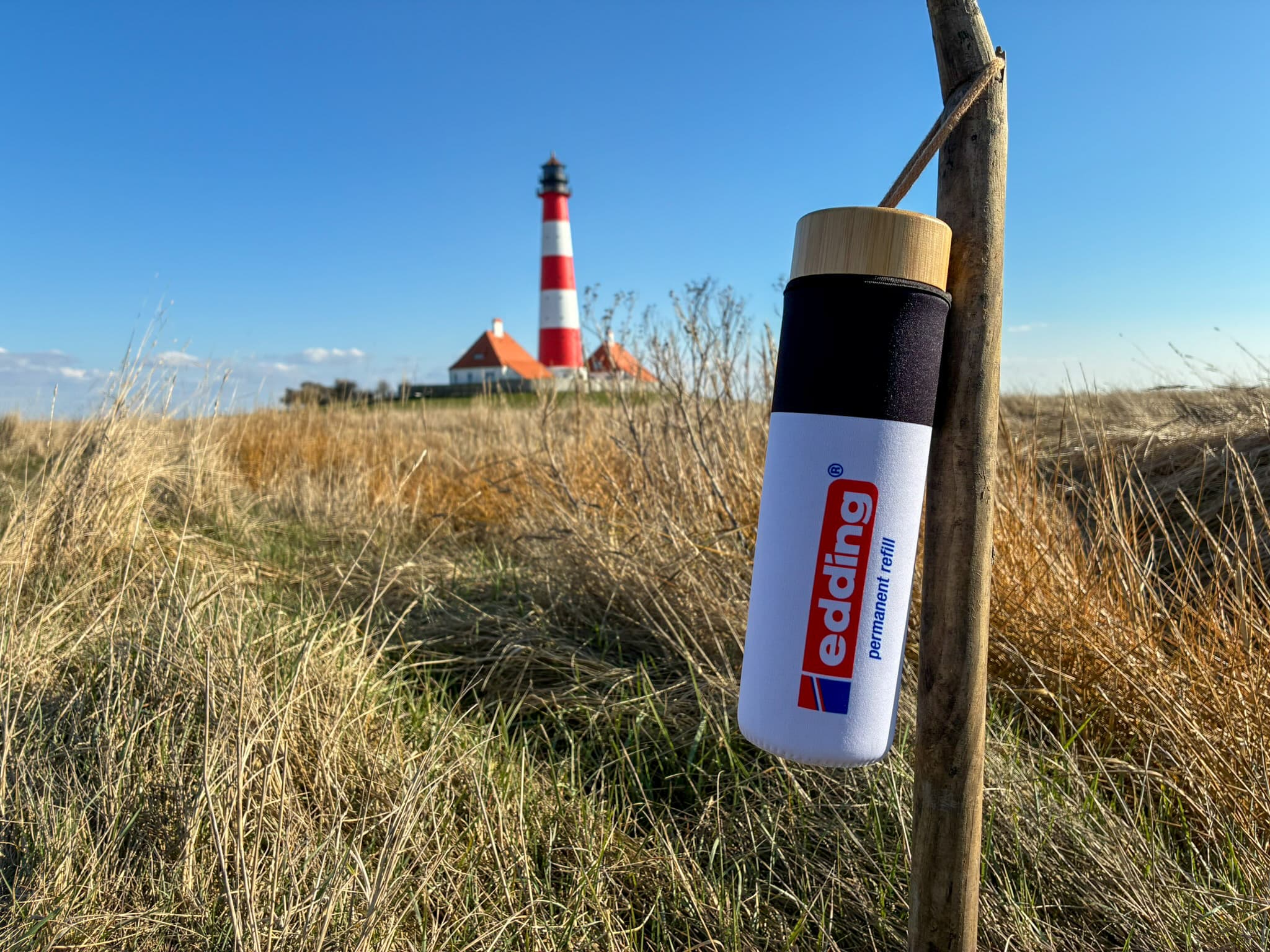 Eine edding Glasflasche mit 650 ml Fassungsvermögen steht inmitten einer Wiese, im Hintergrund ragt stolz unter einem wolkenlosen blauen Himmel ein rot-weiß gestreifter Leuchtturm empor.