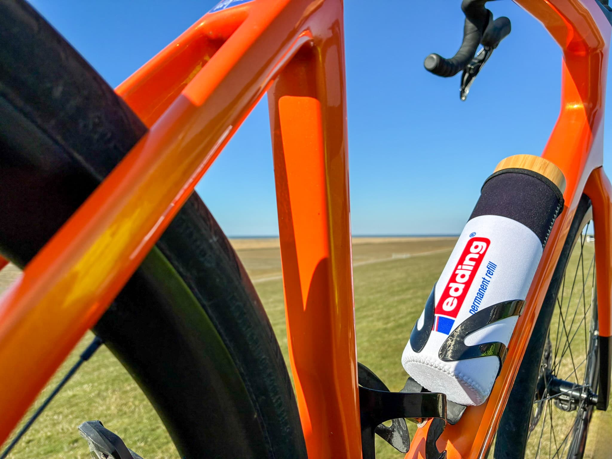 Nahaufnahme eines orangefarbenen Fahrradrahmens mit einer weißen edding-Glasflasche (650 ml) und einem Deckel in Bambusoptik vor einem klaren blauen Himmel und einer weiten Wiese, die Abenteuer und umweltfreundlichen Stil verkörpert.