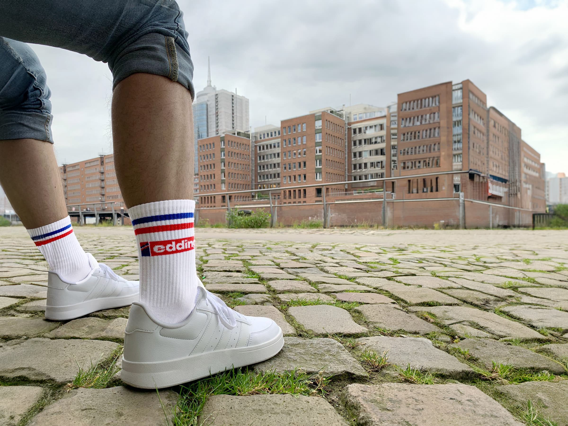Eine Person trägt weiße Turnschuhe und Unisex-Tennissocken von edding mit roten und blauen Streifen und steht auf einer Straße mit Kopfsteinpflaster. Im Hintergrund sind moderne Backsteingebäude und ein bewölkter Himmel zu sehen.