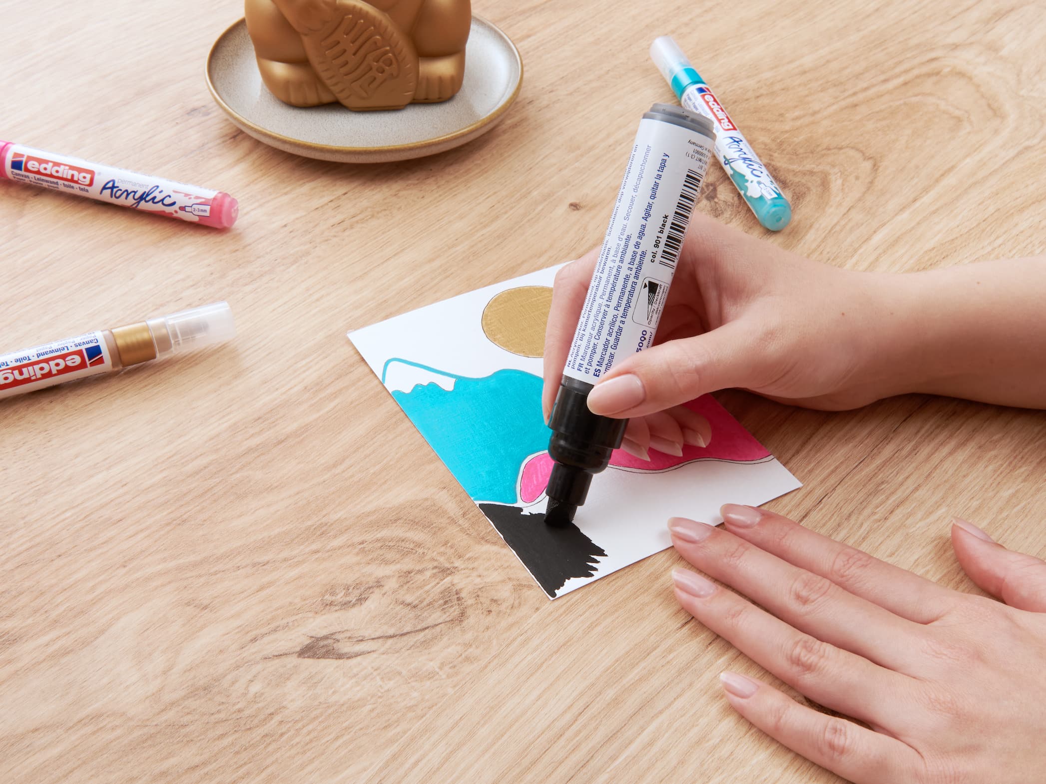 Die Hände einer Person färben mit einem edding 5000 Acrylmarker breit einen Abschnitt eines Papiers mit einem abstrakten Design, zusammen mit anderen Malutensilien wie Markern und einem Stempel auf einem.