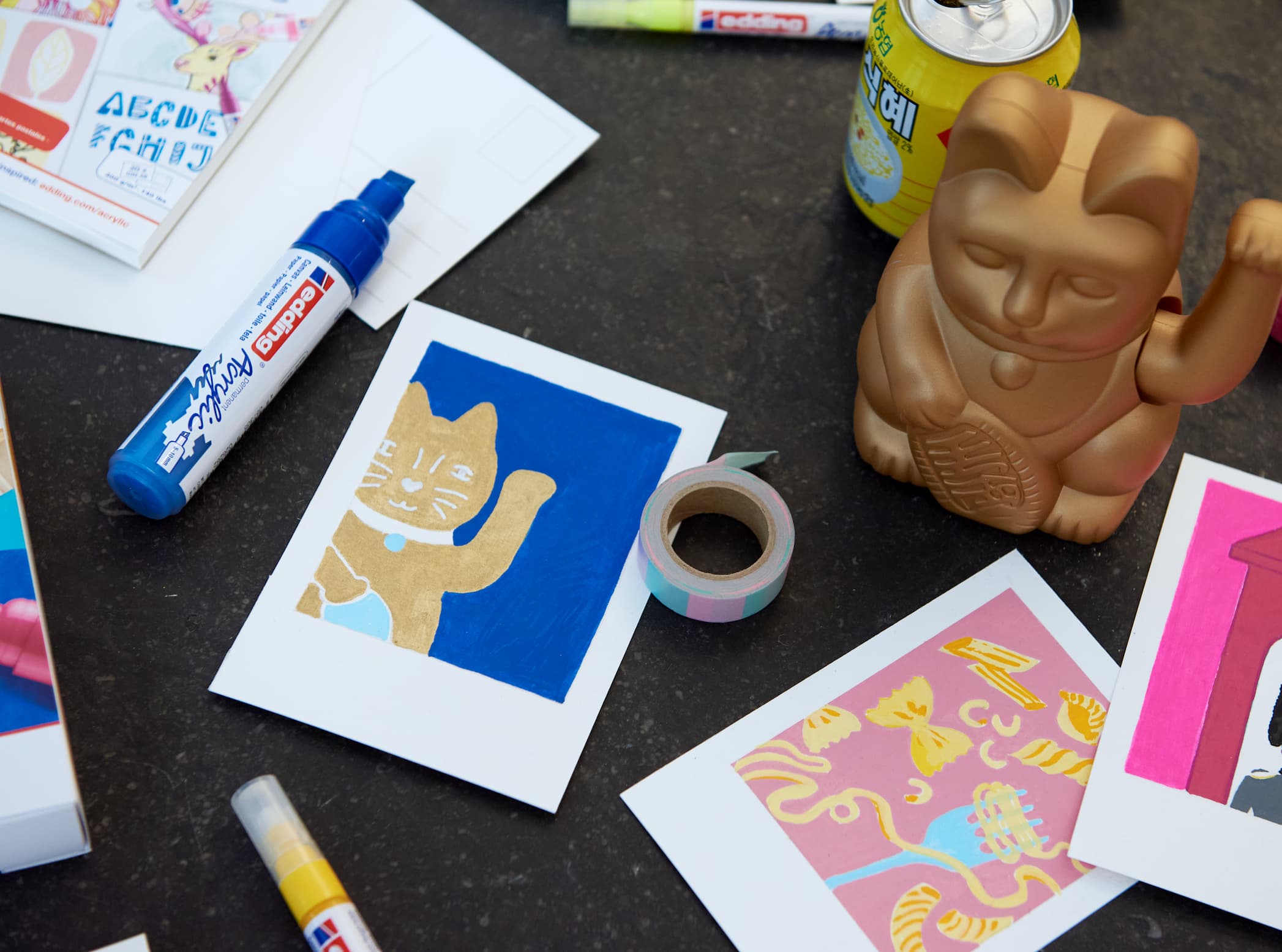 Ein kreativer Arbeitsplatz mit Künstlerbedarf, darunter edding 5000 Acrylmarker breit mit hochpigmentierter Tinte, ausgeschnittene Katzenillustrationen und eine skurrile Affenfigur zwischen buntem Papier auf einer dunklen Oberfläche.