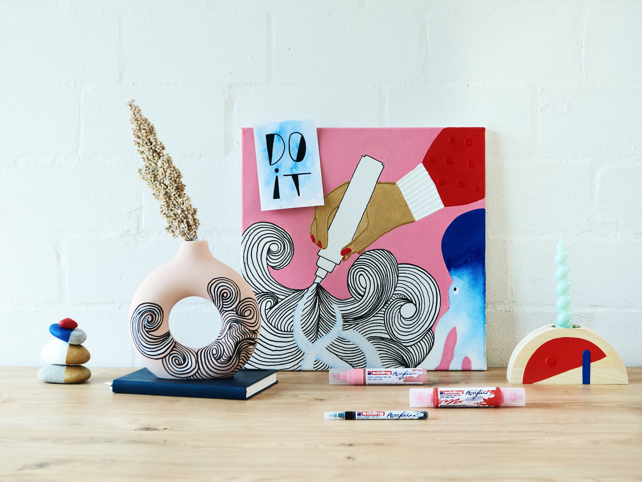 Ein lebendiger und künstlerischer Arbeitsplatz mit einem motivierenden Poster, einem farbenfrohen abstrakten Gemälde, vielseitigen Schreibtischornamenten und einer Auswahl von edding 5000 Acrylmarker breit mit hochpigmentierter Tinte auf einem weißen Holztisch davor