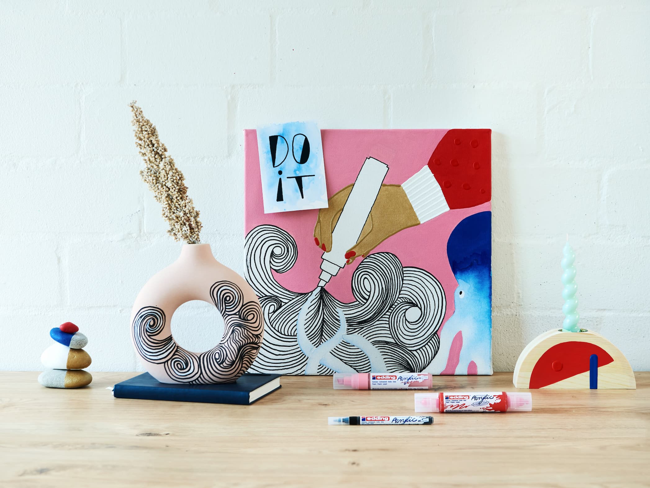 Ein kreativer und farbenfroher Arbeitsplatz mit künstlerischen Postern, einer Vielzahl einzigartiger Dekorationsgegenstände und edding 5100 Acrylmarker-Mediumzubehör auf einem Holztisch vor einer weißen Ziegelwand.