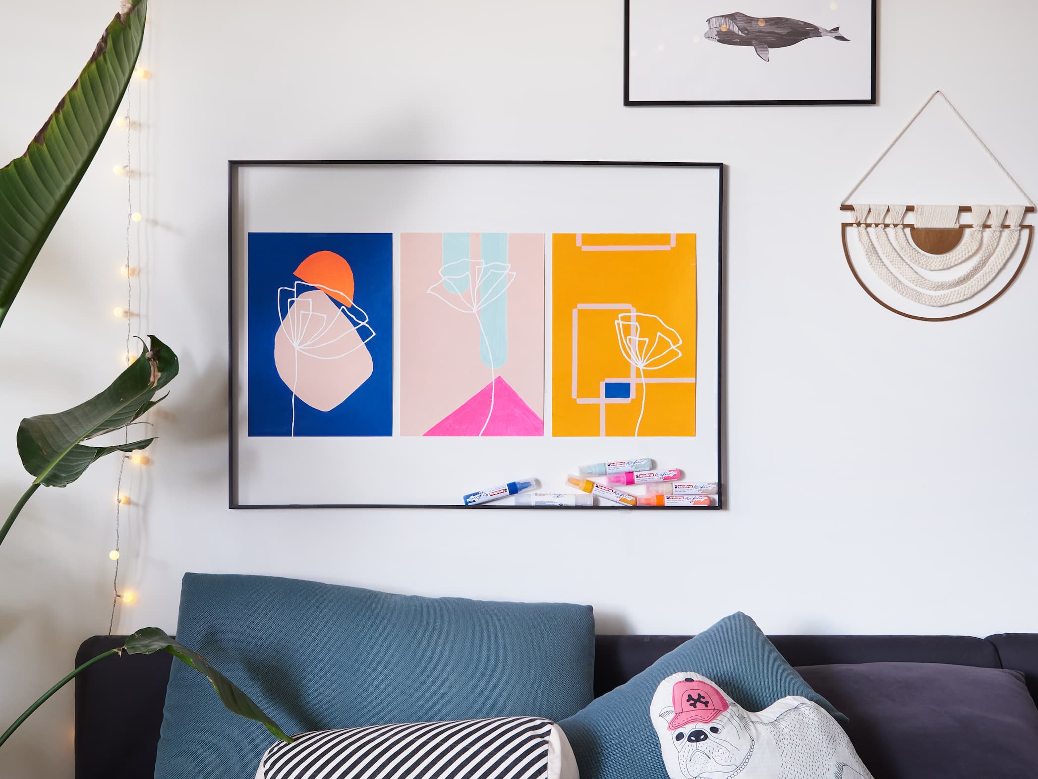 Moderne Wohnzimmerecke mit einem farbenfrohen abstrakten Kunstwerk, erstellt mit dem edding 5000 Acrylmarker breit von edding über einem blauen Sofa mit dekorativen Kissen, ergänzt durch eine Lichterkette, Haus.