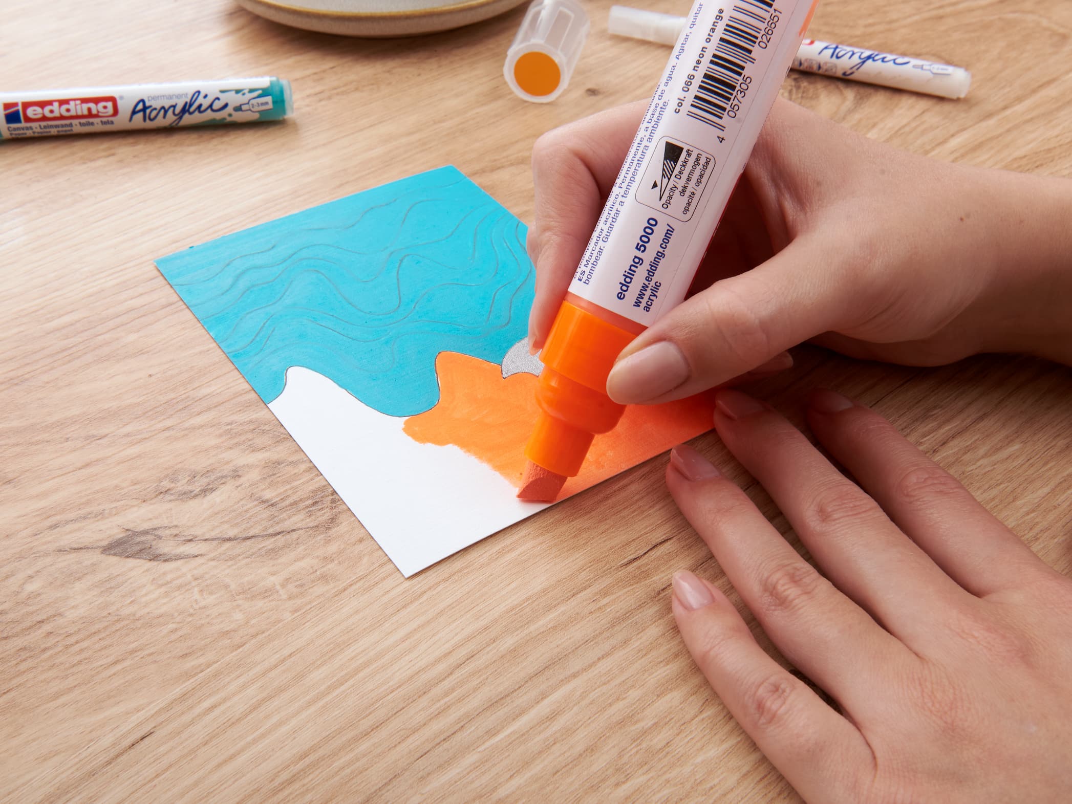 Eine Person hält einen edding 5000 Acrylmarker breit in der Hand und fügt einem abstrakten blauen und orangefarbenen Kunstwerk auf einem Holztisch eine lebendige Ebene hinzu.