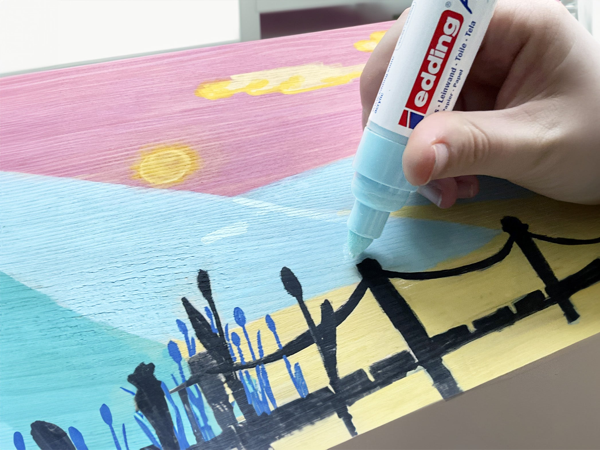 Eine Person, die mit einem edding 5000 Acrylmarker breit Details zu einem farbenfrohen Gemälde eines Sonnenuntergangs hinzufügt.