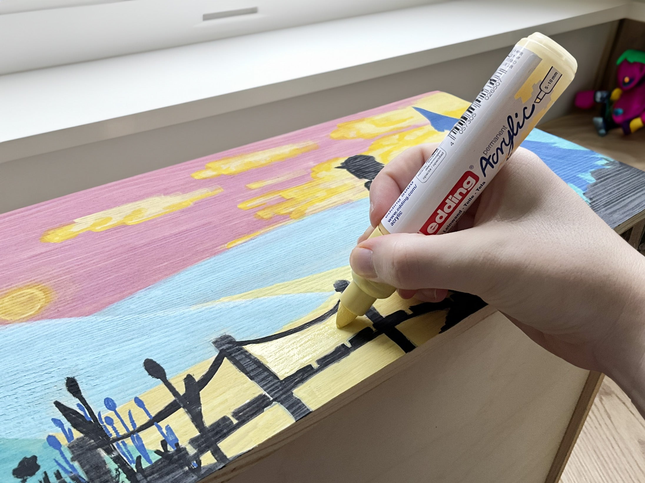 Die Hand eines Künstlers hält einen gelben edding 5000 Acrylmarker breit und fügt Details zu einem farbenfrohen Leinwandgemälde hinzu.