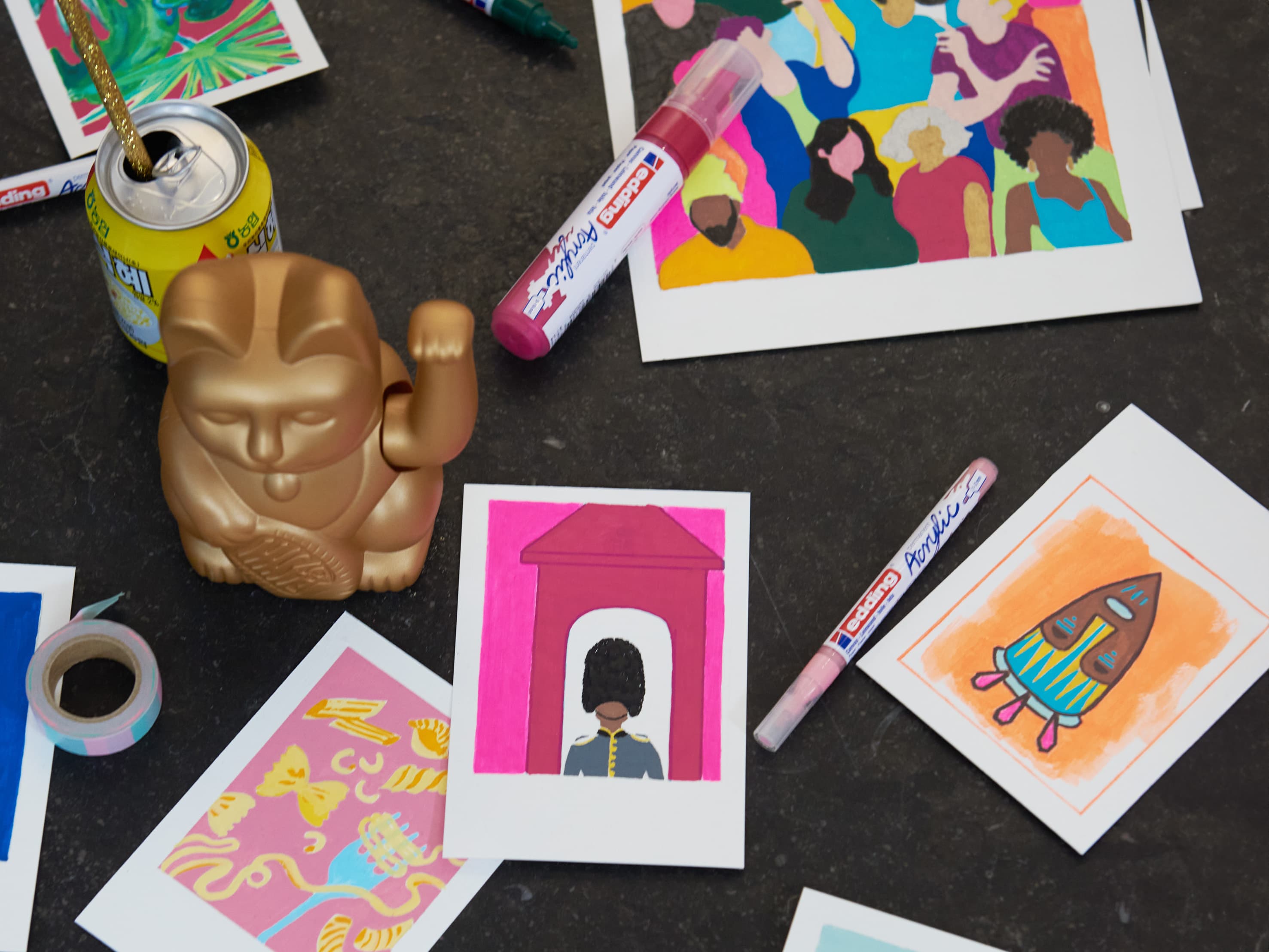 Ein lebendiger und farbenfroher Künstlerarbeitsplatz mit einer Auswahl an Illustrationen zu unterschiedlichen Themen, flankiert von Markern, einem edding 5000 Acrylmarker breit und einer skurrilen Goldfigur.
