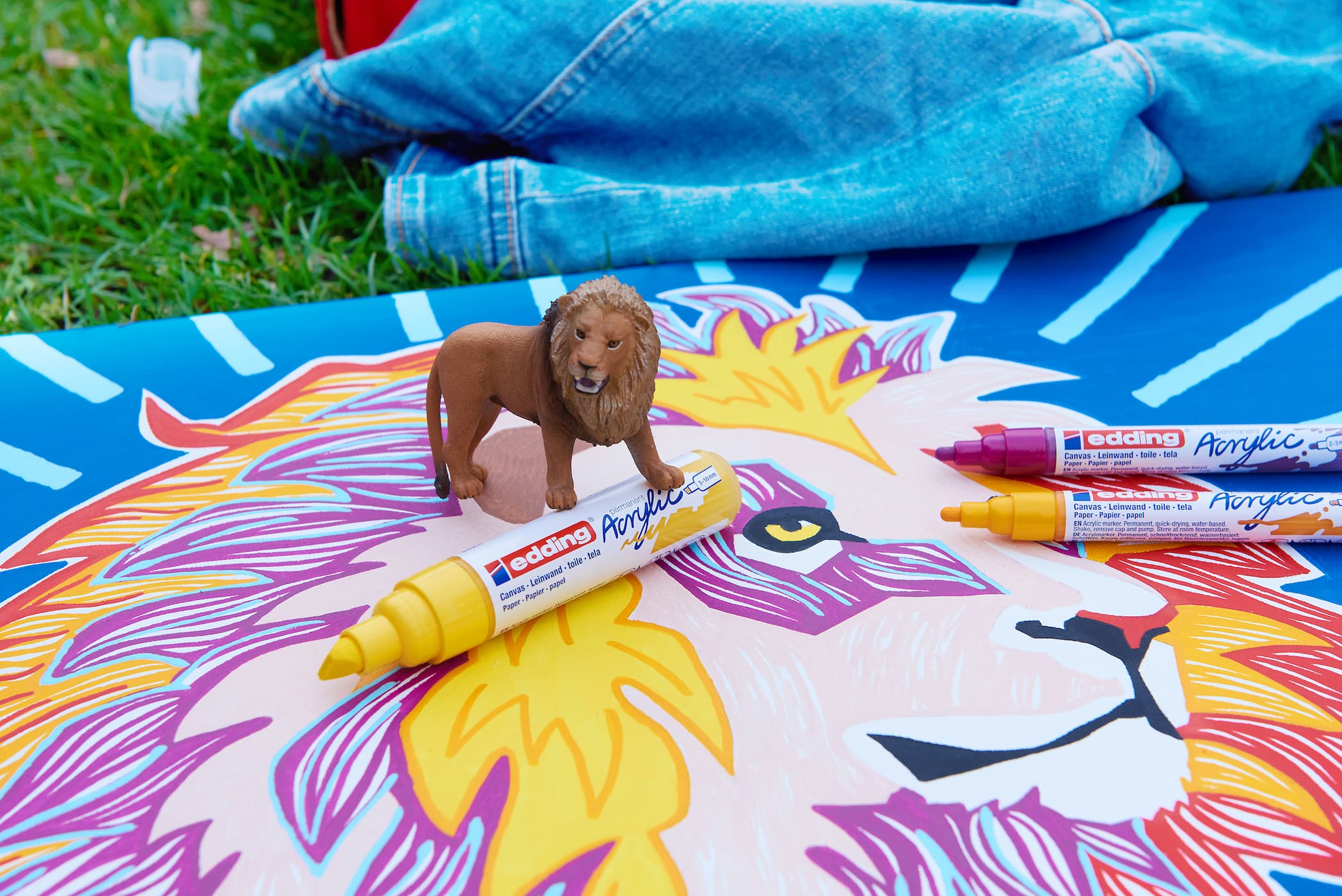 Ein kleiner Spielzeuglöwe steht auf einer großen, farbenfrohen Zeichnung eines Löwengesichts, begleitet von einer Auswahl an bunten Markern, darunter der edding 5000 Acrylmarker breit mit Hochpig.