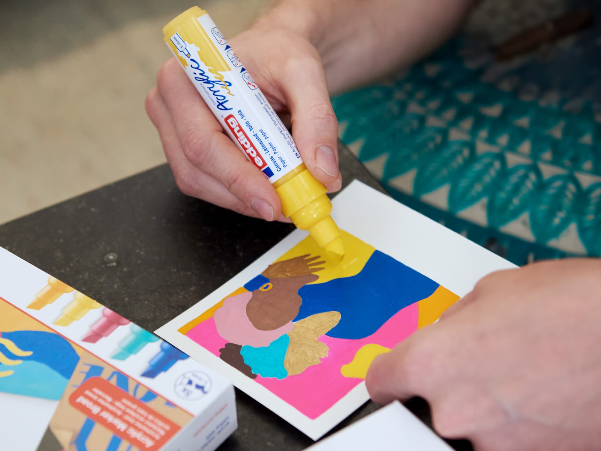 Die Hände einer Person tragen den edding 5000 Acrylmarker breit von edding auf ein farbenfrohes ausgeschnittenes Kunstwerk auf, das mit hochpigmentierten Tinte-Details verziert ist, auf einem Tisch.